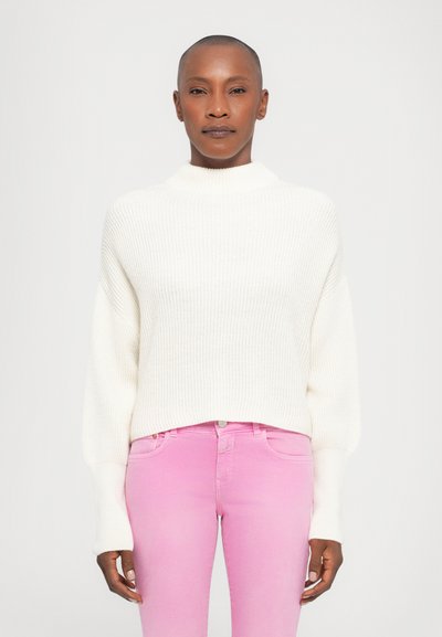 Pull en tricot blanc épais avec col montant et manches longues effilées, associé à un jean rose ajusté. Texture lisse et coupe décontractée.