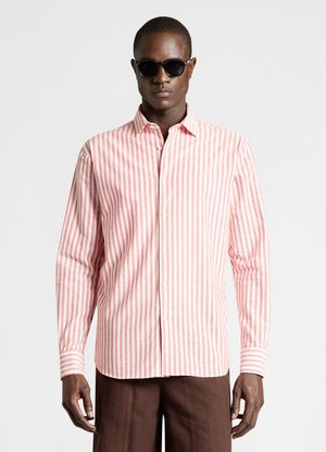 Man wearing black sunglasses, a pink and white vertically striped button-up shirt, and brown pants, standing against a plain background.