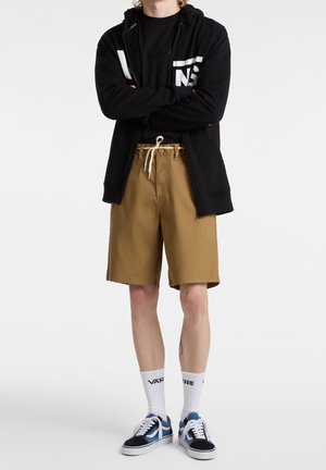 Sudadera con capucha negra con cremallera, camiseta negra, pantalones cortos caqui, calcetines blancos con logo, zapatillas azules y negras. Conjunto informal con un ajuste cómodo.