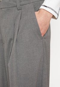 Gray tailored trousers made of textured fabric, featuring side pockets and sharp pleats, shown with a hand resting in a pocket.