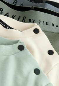 Drei T-Shirts, die in einem Stapel präsentiert werden: eines in hellem Grün, eines in Creme und eines in hellem Grau. Jedes hat schwarze Knopfakzente und einen weichen Stoff.