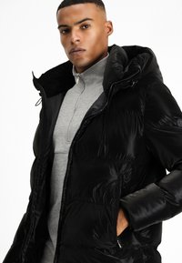 Chaqueta acolchada negra con capucha, con una textura suave, bolsillos con cremallera y un diseño ajustado sobre una cima de cuello alto gris.