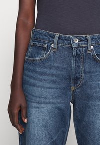 Niebieskie dżinsy z pośrednim stanem, pięcioma kieszeniami i pomarańczowymi przeszyciami. Materiał jest wytrzymały z delikatnym efektem sprania na tkaninie.