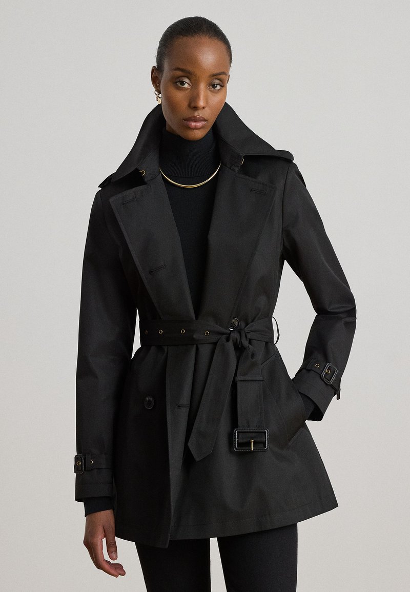 Vrouw draagt een zwarte trenchcoat met ceintuur over een zwarte coltrui, geaccentueerd met een gouden ketting en oorbellen, staand voor een effen achtergrond.