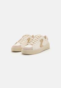 Sneakers beige a bassa altezza con pannelli laterali testurizzati, lacci e suole spesse, mostrati come un paio inclinato a sinistra su uno sfondo bianco.