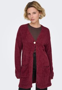 Vinrød cardigan i blødt, pelsagtigt materiale; åben front med en dekorativ blomstknap, to forlommer og lange ærmer.