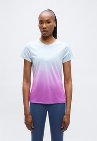 Γυναίκα με κοντομάνικη αθλητική μπλούζα σε διαβάθμιση από ανοιχτό μπλε σε μωβ και σκούρο μπλε κολάν, στέκεται μπροστά σε απλό λευκό φόντο.