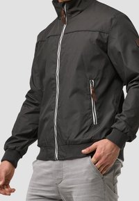 Blouson aviateur noir avec un col montant, une fermeture éclair frontale et deux poches latérales. Présente des accents de fermeture éclair blancs et des languettes de tirage en cuir marron.