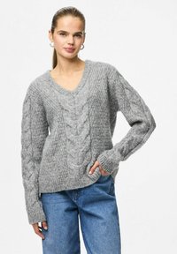 Femme aux longs cheveux en queue de cheval portant un pull gris en maille torsadée avec un col en V et un jean bleu, se tenant avec une main dans la poche.