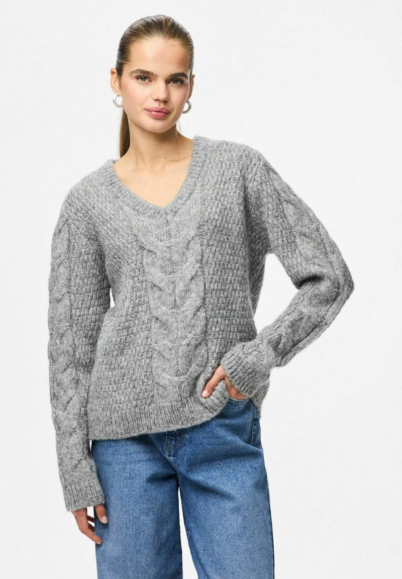 Femme aux longs cheveux en queue de cheval portant un pull gris en maille torsadée avec un col en V et un jean bleu, se tenant avec une main dans la poche.