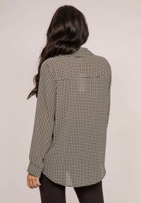 Chemise à motif pied-de-poule noir et blanc avec des manches longues, un col et un ourlet légèrement courbé. Détails de boutons sur les poignets visibles.