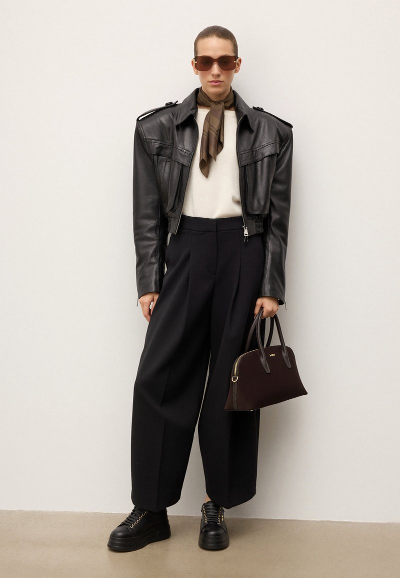 Veste en cuir noire courte, pull crème, pantalon large noir, sac à main marron, baskets noires avec accents dorés, lunettes de soleil, écharpe marron.