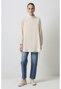 Tunique crème à manches longues avec un col haut et des cordons de serrage réglables au niveau de l'ourlet, associée à un jean bleu à jambe droite et à des baskets beiges.