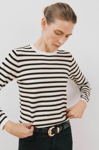 Gestreiftes Langarmoberteil in Schwarz, Weiß und Gold. Rippkragen und -bündchen. Modell justiert den Saum bei getragenen schwarzen Hosen und einem Gürtel mit goldener Schnalle.