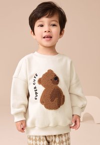 Kermansävyinen collegepaita, jossa on kuvioitu ruskea karhuhöyhen ja teksti "SUPER CUTE". Resorit hihoissa ja helmassa. Yhdistetty kuvioituihin beigenvärisiin housuihin.