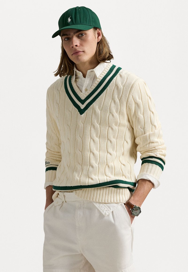 Creme kabelstrik sweater med grønne v-hals striber, parret med en grøn kasket og creme shorts. Bemærkelsesværdigt ur på håndleddet med læderrem.