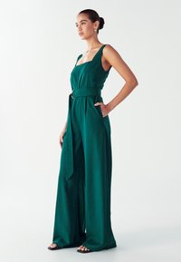 Mujer en un mono verde sin mangas con pantalones anchos y cinturón, de pie con una mano en el bolsillo frente a un fondo blanco liso.