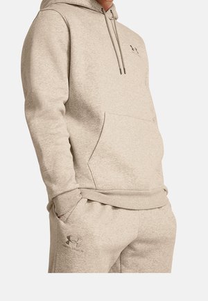 Beżowy komplet Under Armour z bluzą z kapturem i dresowymi spodniami z przednią kieszenią, logo na klatce piersiowej i lewym udzie, noszony przez osobę z rękami w kieszeniach.