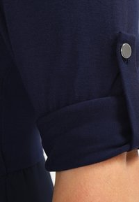 Haut bleu marine à manches courtes avec poignets retroussés, en tissu doux. Présente un bouton argenté sur le poignet pour plus de détails.