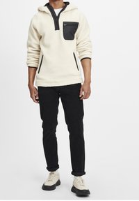 Fleece hoodie in crème met zwarte accenten, kwart-rits, voorzak en steekzakken, gecombineerd met zwarte broek en lichte sneakers.