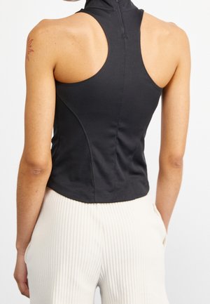 Achteraanzicht van een persoon die een mouwloze zwarte coltrui met rits en witte geribde broek draagt.