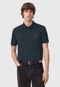Dunkelblauses Poloshirt mit Kragen, dreiknöpfiger Knopfleiste und kleinem gesticktem Logo. Getragen mit schwarzen Jeans und einem schwarzen Gürtel.