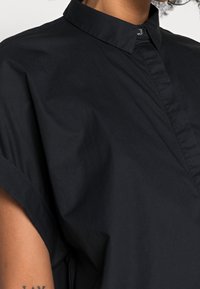 Camicia nera in cotone con colletto a punta, maniche corte e una placca con un singolo bottone. Tessuto liscio con una vestibilità rilassata.