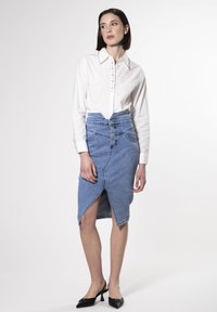 Jupe crayon en jean à taille haute, avec un devant à cinq boutons et une fente jusqu'à la cuisse ; associée à une chemise blanche à boutons et des escarpins noirs.