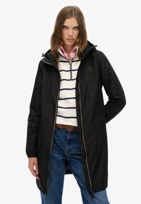 Veste imperméable noire avec zip à l'avant et capuche. Comprend un logo sur la poitrine, associée à une chemise rayée et un jean bleu.