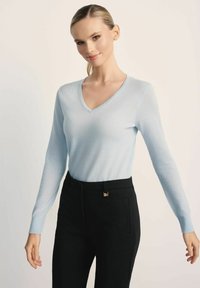 Jasnoniebieski sweter z dekoltem w serek i długim rękawem, uszyty z miękkiej tkaniny, w połączeniu z czarnymi spodniami o dopasowanym kroju, ozdobionymi złotym akcentem w talii.