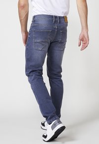 Pantalones vaqueros de mezclilla azul oscuro con corte ajustado, que presentan múltiples bolsillos y detalles de costura. Se llevan con zapatillas en blanco y negro.
