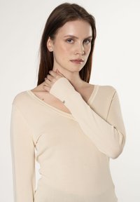 Beige Langarmoberteil mit V-Ausschnitt und figurbetontem Design. Mit gerippten Details und einem kleinen Akzent am Handgelenk. Weiche, glatte Textur.