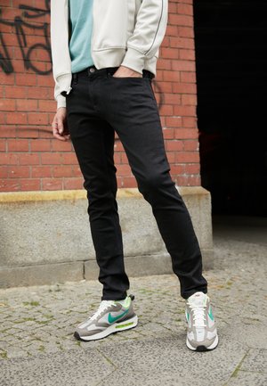 Personne portant un jean slim noir, des baskets blanches et turquoises, debout contre un mur en briques rouges sur un trottoir pavé.