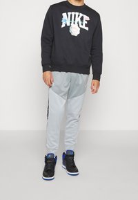 Persona con sudadera negra de Nike, pantalones de chándal gris claro de Nike con franja lateral y zapatillas de alta de Nike en negro y azul, de pie sobre un suelo claro.