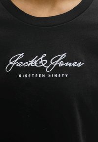 Melnā kreklu ar apaļu kakla izgriezumu, ar baltu izšūtu "Jack & Jones" logotipu un "Nineteen Ninety" tekstu centrā uz krūtīm.