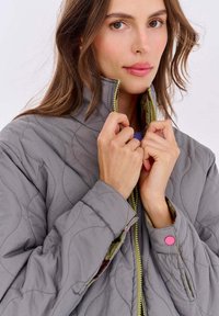 Prešívaná sivá bunda s vysokým golierom, s zeleným zipsom, ružovými cvokmi na rukávoch a textúrovanou tkaninou.