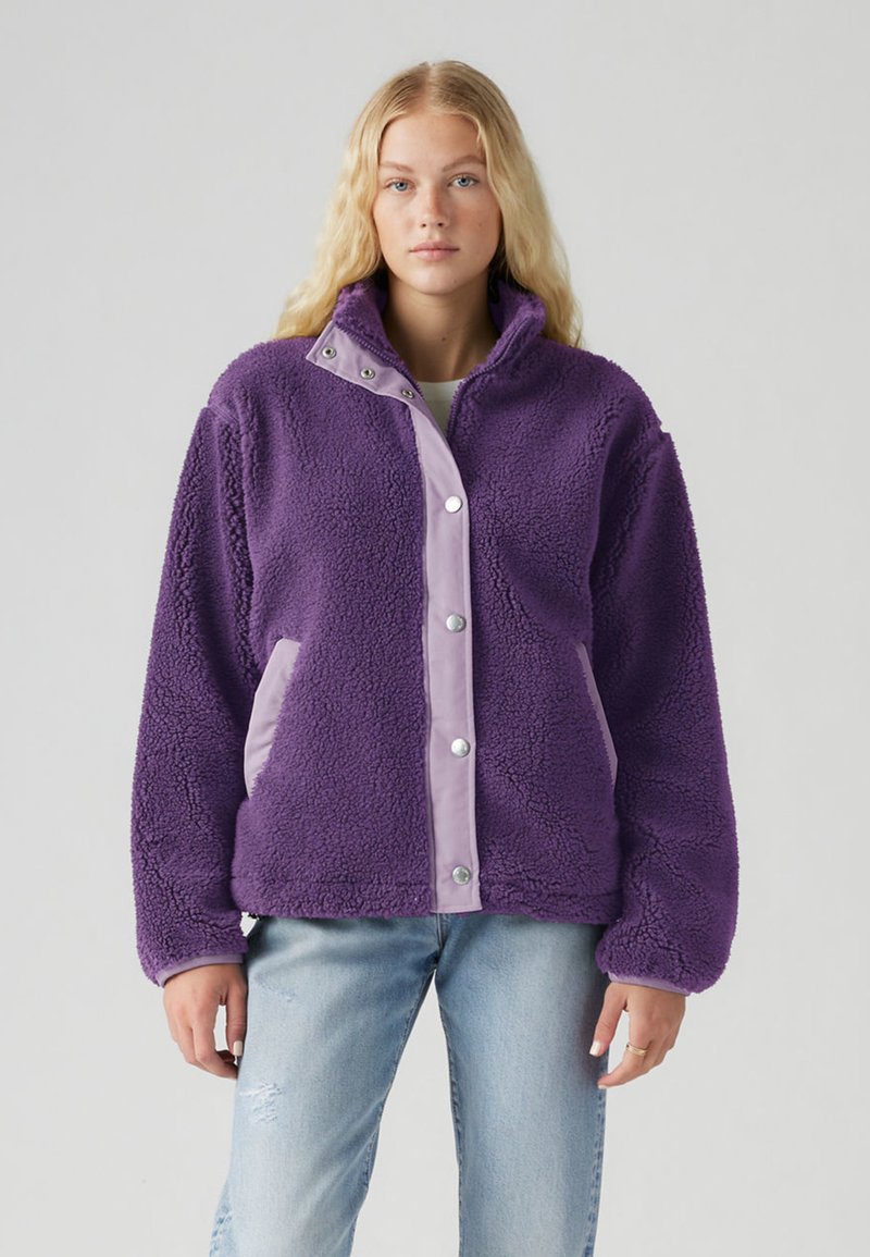 Veste polaire violette avec col montant, fermeture à boutons-pression, et accents violet clair sur les côtés. Texture douce et coupe décontractée.