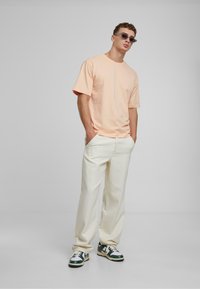 Pfirsichfarbenes Baumwoll-T-Shirt mit kurzen Ärmeln, kombiniert mit beigefarbenen, lässigen Hosen. Die Schuhe sind grüne und weiße Sneakers mit detaillierten Akzenten.