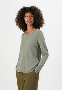 Donna con capelli ricci che indossa una maglietta a maniche lunghe di un verde chiaro e pantaloni marroni, in piedi con una mano in tasca, mentre guarda di lato.