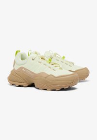 Zapatillas Lacoste en una combinación de colores beige claro y crema, con parte superior de malla, suela texturizada y acentos en verde brillante, presentando un cierre con cordones.