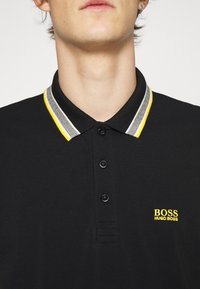 Homme portant un polo noir avec un col rayé blanc et jaune et un logo jaune Hugo Boss sur la poitrine.