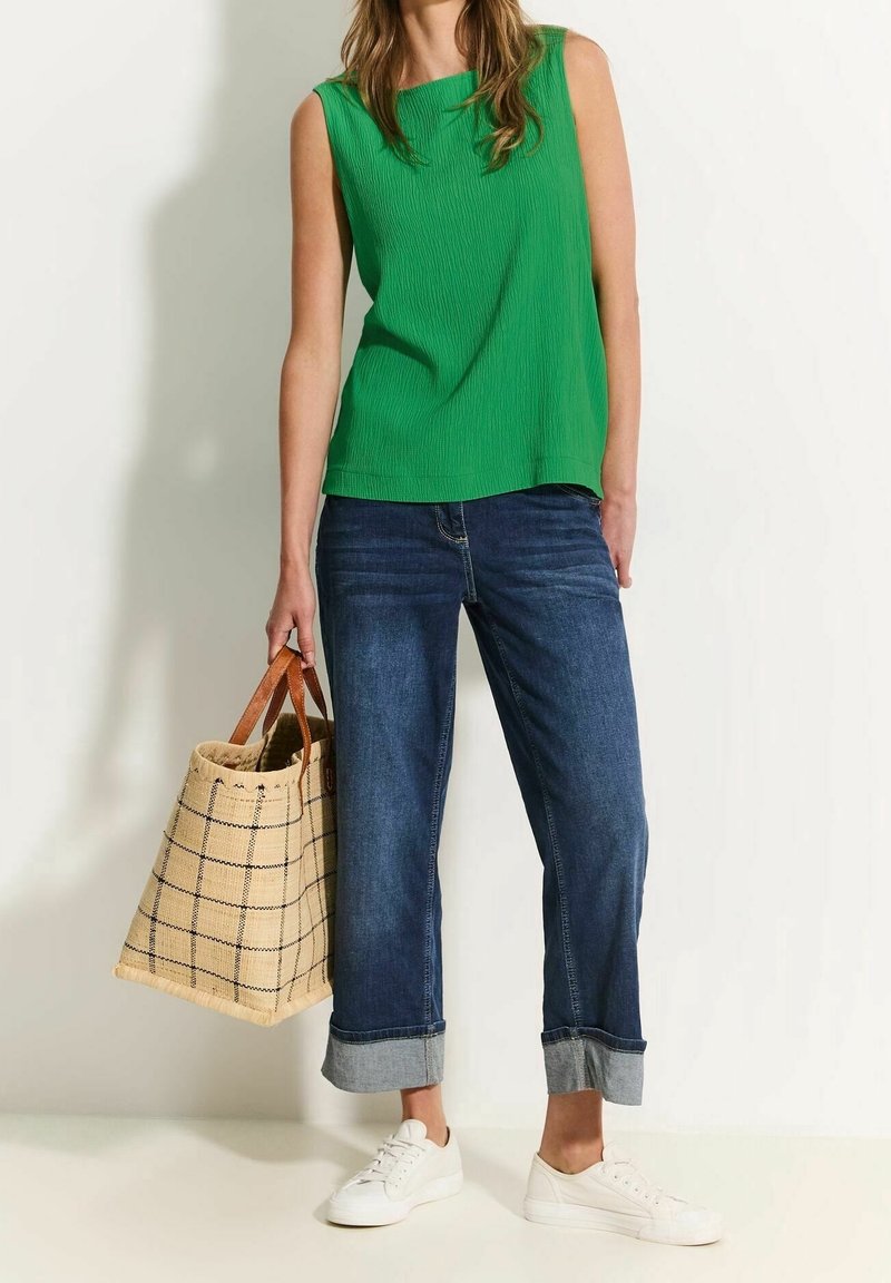Grünes, geripptes, ärmelloses Top, blaue, umgekrempelte Jeans, weiße Sneakers und eine beigefarbene Tragetasche mit Karomuster und braunen Ledergriffen.