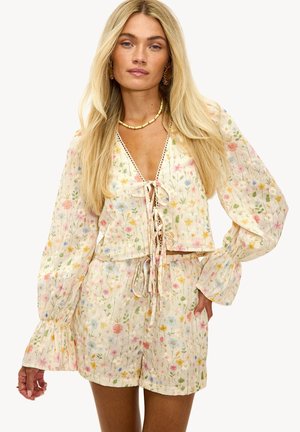 Vrouw die een crèmekleurige lange mouw blouse met bloemenprint en strikjes draagt, gecombineerd met bijpassende shorts met hoge taille, gecompleteerd met een gouden ketting en oorbellen.