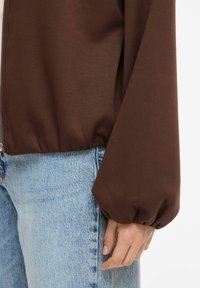 Pruun, kerge jakk, millel on tõmblukuga sulgemine, padjandiga varrukad ja elastsed mansetid, kantud hele siniste teksapükste peal. Sile tekstuur.