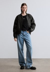Veste aviateur en cuir noir, jean large décontracté en denim bleu, chaussures compensées noires, avec un T-shirt noir ajusté portant un texte blanc.
