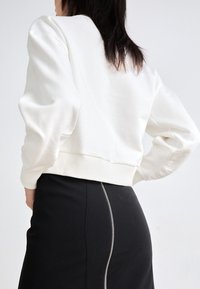 Mujer vistiendo una blusa blanca de manga larga y una falda negra con una cremallera visible que baja por la parte posterior, de pie con las manos en las caderas.