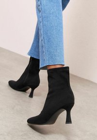 Czarne zamszowe botki za kostkę z ostrymi noskami i 6 cm obcasami typu stiletto, zestawione z niebieskimi, skróconymi dżinsami, prezentujące smukły design.