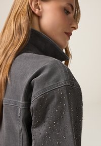 Giacca di jeans grigia con colletto alto, realizzata in un tessuto texturizzato e con applicazioni di borchie argentate sulle spalle.