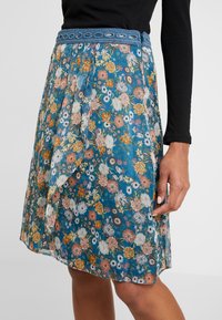Blomsteret nederdel med blå baggrund, prydet med farverige blomster i orange, pink og hvid. Blondekant i taljen med et plisseret design.