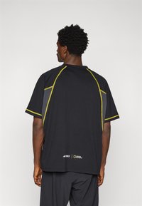 T-shirt atlético preto com mangas curtas, costuras amarelas e detalhes cinzentos. Apresenta uma marca perto da bainha. A textura parece suave e leve.